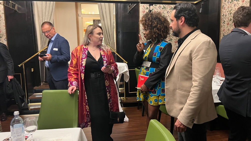 Drei Personen im Gespräch miteinander, eine Person stützt Hände an grünem Stuhl ab, im Hintergrund floral gemusterte Tapete, Holzvertäfelung und weitere Personen im Gespräch