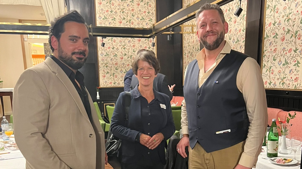 Drei lächelnde Personen nebeneinander stehend, im Hintergrund floral gemusterte Tapete mit Holzvertäfelung