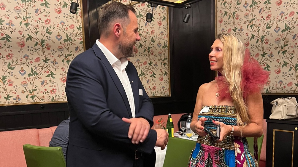 Zwei lächelnde Personen in Seitenansicht im Gespräch miteinander, im Hintergrund floral gemusterte Tapete und Holzvertäfelung