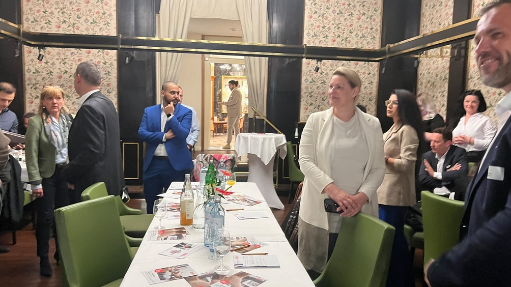 Mehrere Personen in Raum stehend, teilweise in Gesprächen miteinander, zentral ein Tisch mit Prospekten und Getränken umgeben von grünen Stühlen