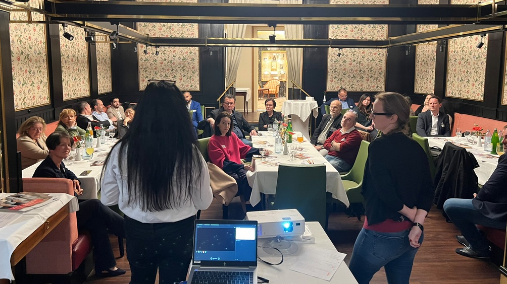 Zwei Personen in Rückenansicht vor Laptop und auf grünen Stühlen an Tischen sitzendem Publikum sprechend