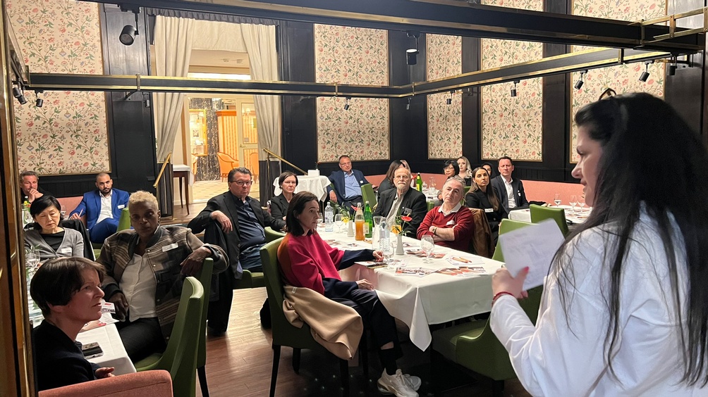 Person in Seitenansicht vor auf grünen Stühlen an Tischen sitzendem Publikum sprechend