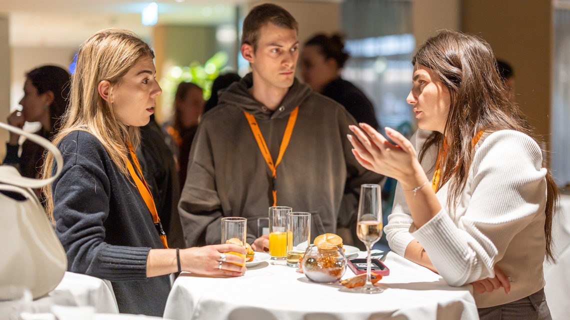 Zu sehen sind Personen, die an einem runden Tisch stehen. Auf diesem sind Gläser und Snacks  platziert. 