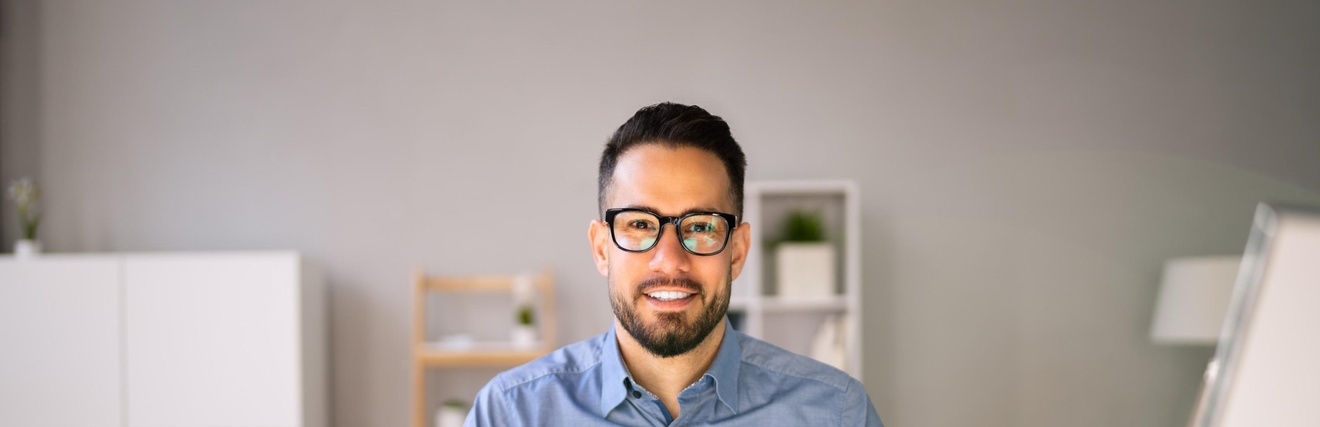 Lächelnde Person mit Brille sitzt an einem Schreibtisch und arbeitet mit aufgeschlagener Mappe, Taschenrechner, im Hintergrund befinden sich Regale und Ablagen