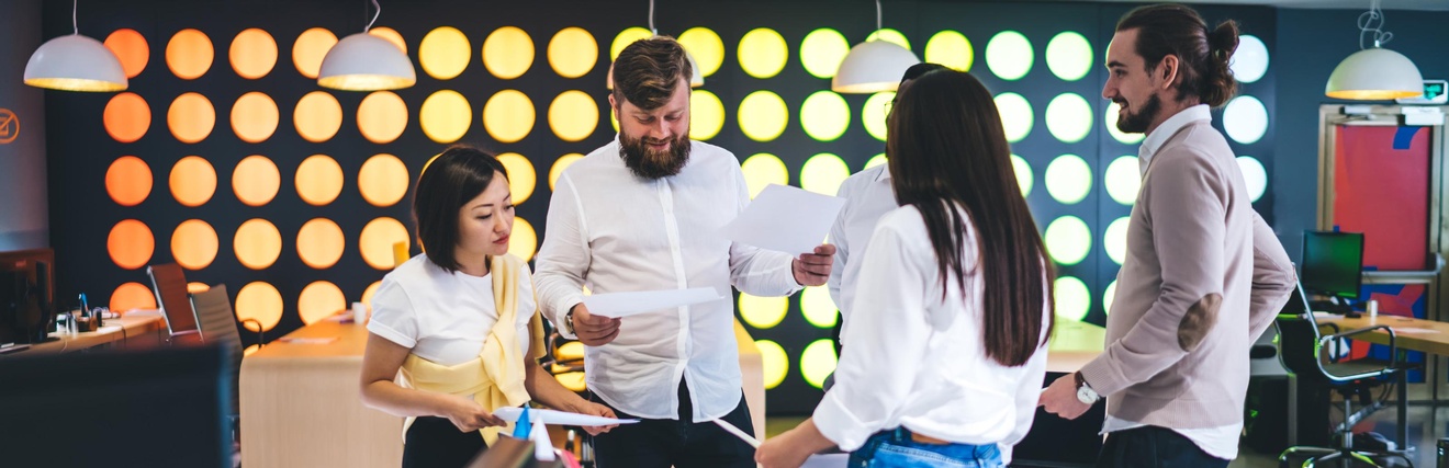 Fünf Personen stehen in einem Büro um einen Tisch. Zwei der Personen halten jeweils einen Zettel. Auf dem Tisch liegen ein Tablet, Post-its, ein Laptop sowie andere Büroutensilien. Die Personen reden miteinander