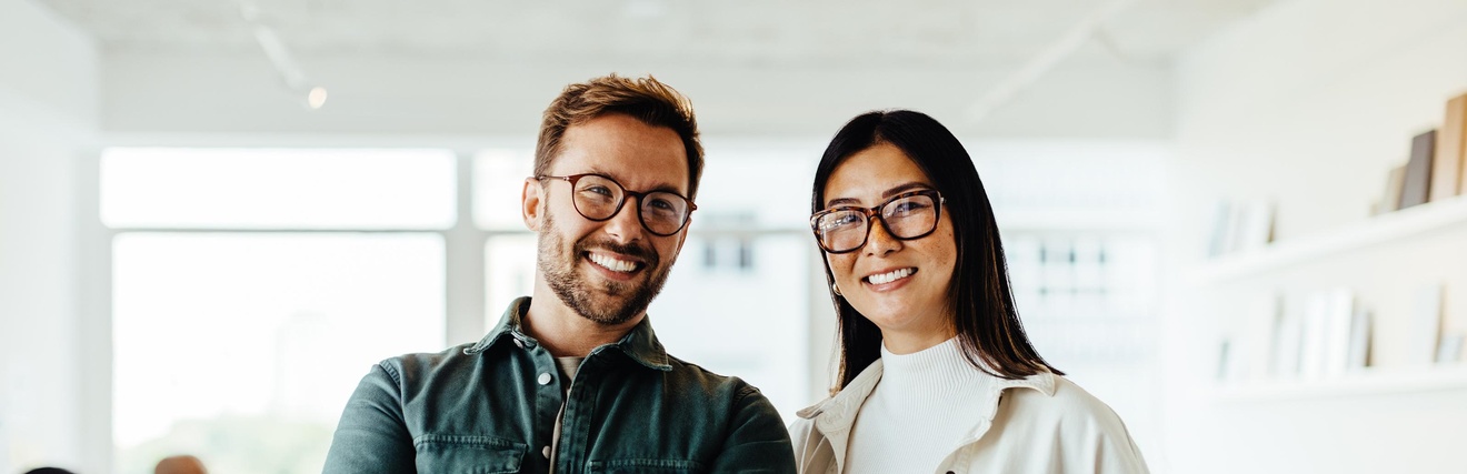 Bild mit 2 Personen, die in einem Büro stehen und in Richtung Kamera blicken. Sie lächeln.
