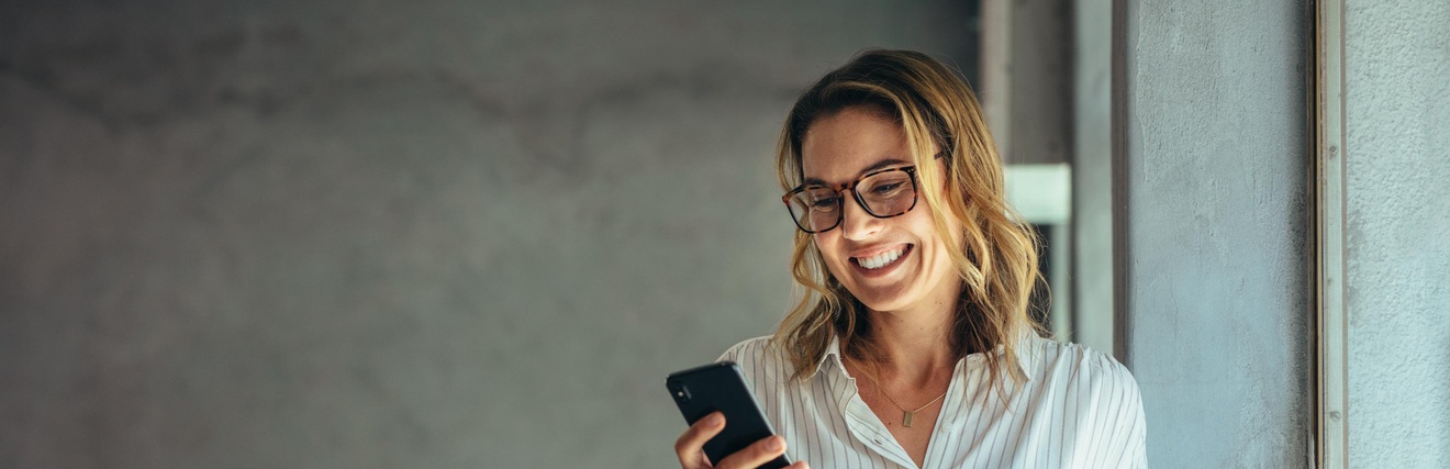 Person mit Brillen blickt freudestrahlend auf Smartphone, im Hintergrund verschwommen Schreibtisch mit Sessel