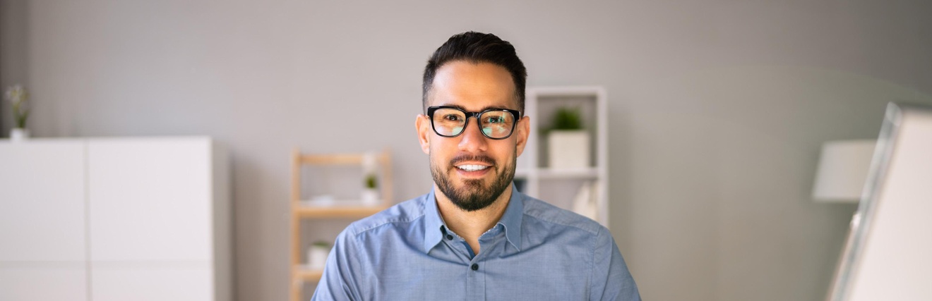 Lächelnde Person mit Brille sitzt an einem Schreibtisch und arbeitet mit aufgeschlagener Mappe, Taschenrechner, im Hintergrund befinden sich Regale und Ablagen