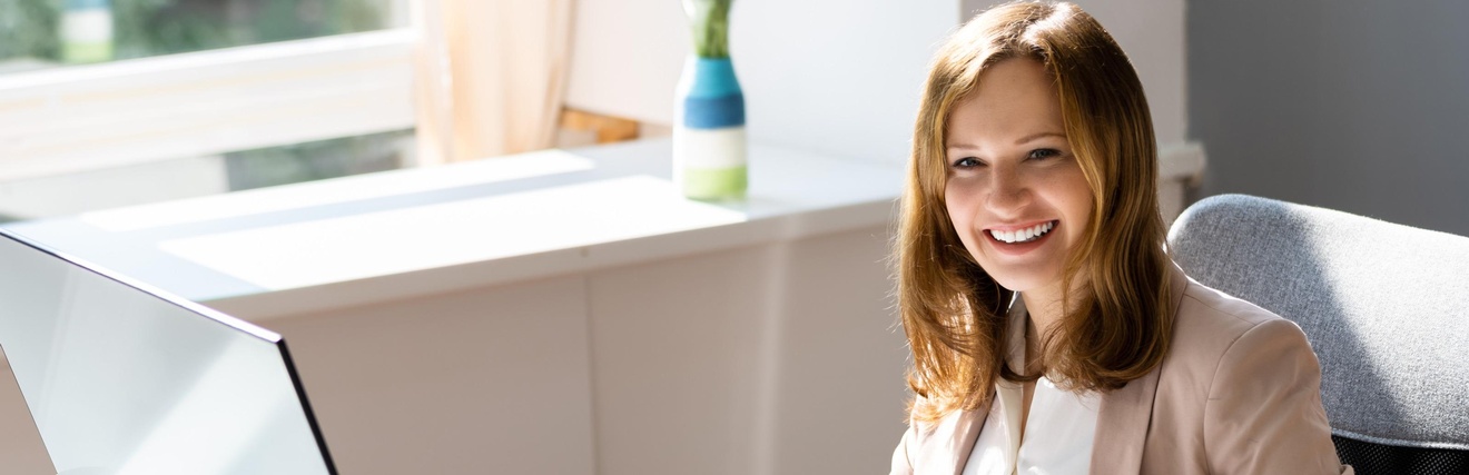 Lächelnde Person sitzt an einem Schreibtisch mit Laptop und geht eine Mappe mit Unterlagen durch, während mit einem Taschenrechner gerechnet wird