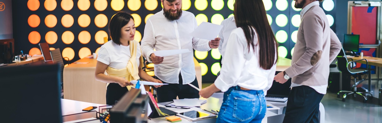 Fünf Personen stehen in einem Büro um einen Tisch. Zwei der Personen halten jeweils einen Zettel. Auf dem Tisch liegen ein Tablet, Post-its, ein Laptop sowie andere Büroutensilien. Die Personen reden miteinander