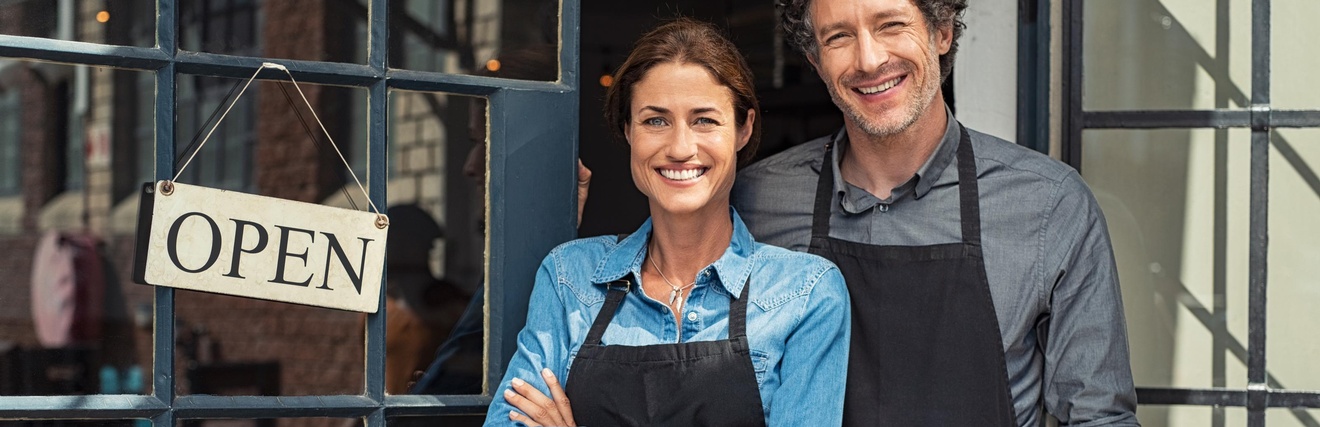 Zwei lächelnde Personen mit schwarzen schürzen stehen im Eingangsbereichs eines Geschäftes, an der Tür hängt ein Schild mit dem Schriftzug open