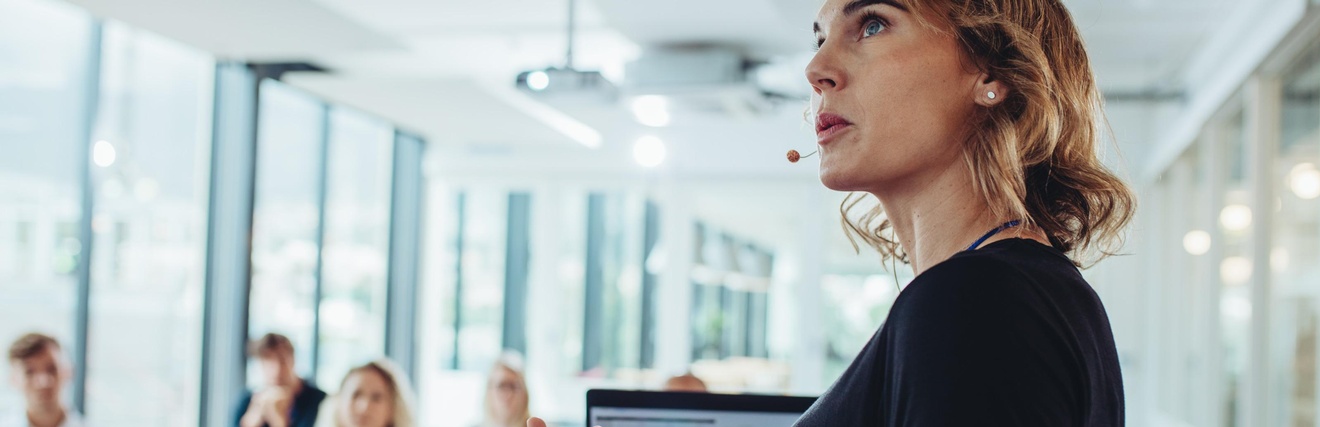 Eine Person mit einem Mikrofon vor den Lippen steht hinter einem Pult und gestikuliert mit den Händen. Auf dem Pult steht ein Laptop. Ihr gegenüber sitzen fünf Personen jeweils einzeln hinter einem Tisch