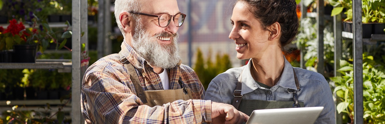 Nahaufnahme von zwei Personen, die in einer Gärtnerei nebeneinander stehen und sich ansehen. Die rechte Person hat einen Bart und eine Brille und deutet auf das Tablet, das die linke Person in den Händen hält. Im Hintergrund Metallregale mit Pflanzen