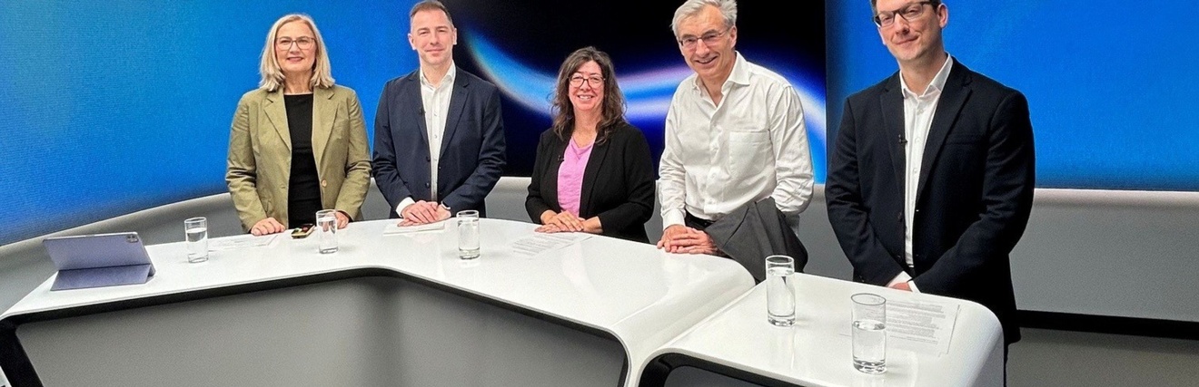Fünf Personen stehen in einem modernen TV-Studio hinter einem Studiotisch.