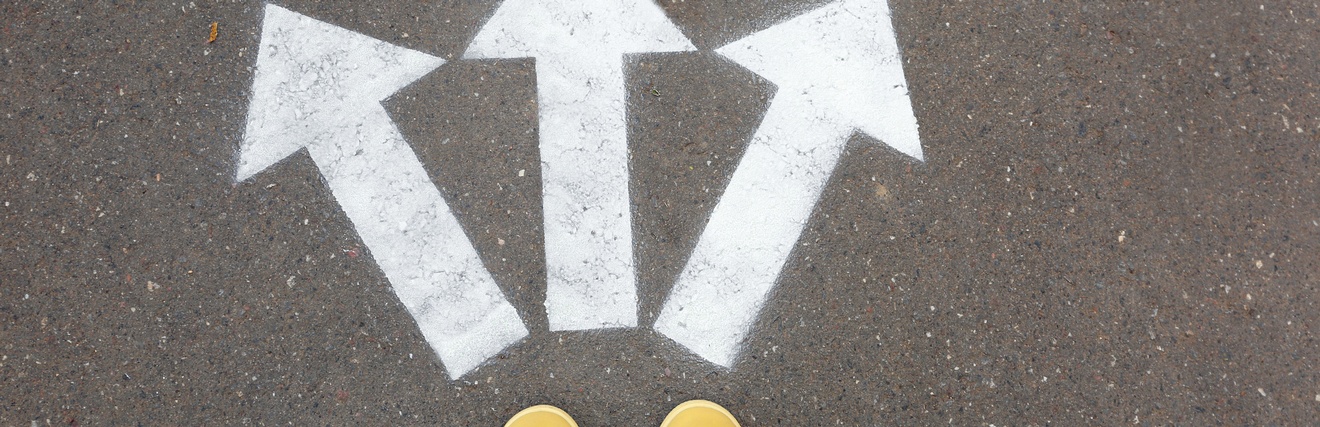 Gelbe Schuhe stehen auf einem Asphalt, davor sind drei weiße Pfeile, die in unterschiedliche Richtungen zeigen am Boden markiert