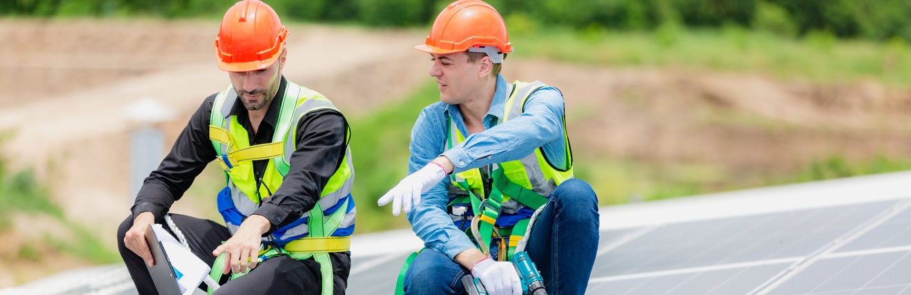 Zwei Personen in gelben Warnwesten mit orangen Schutzhelmen hocken auf Trittfläche auf Dach mit Solarpanelen und deuten auf bestimmte Stelle, im Hintergrund verschwommen belaubt Büsche