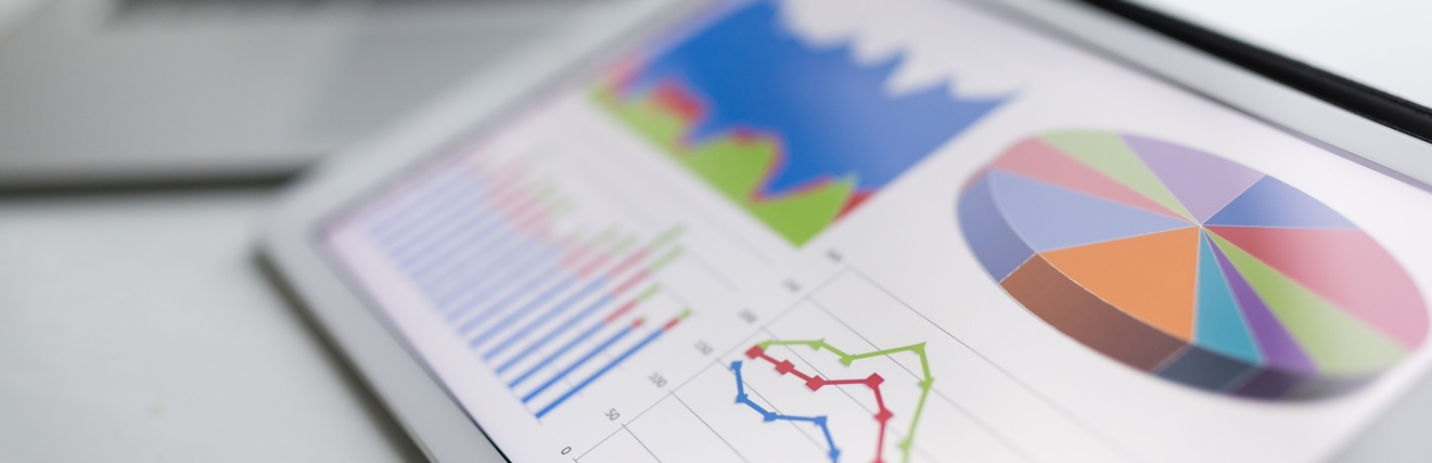 Ansicht eines Tablets mit grafischen Darstellung verschiedener Statistiken. Im Hintergrund ist verschwommen ein Laptop zu sehen.