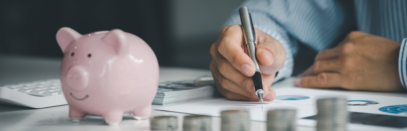 Auf einer Oberfläche sind fünf, unterschiedlich hohe Münzstapel. Links daneben steht eine kleine Figur eines Schweins. Links neben dem Schwein ist ein Taschenrechner. Dahinter sind die Arme einer Person. Unter der rechten Hand sind bedruckte Zettel