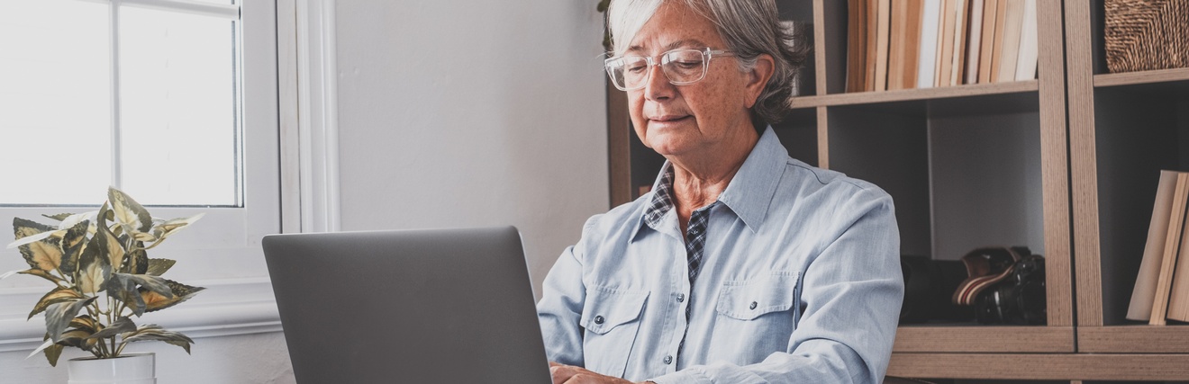 Person mit grauen Haaren und Brille sitzt an Tisch und blickt auf Laptop