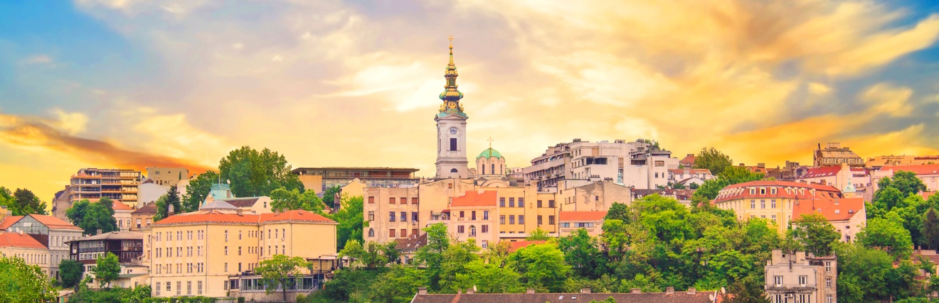 Panoramaaufnahme der Stadt Belgrad mit historischen und modernen Gebäuden. In der Mitte ragt ein Kirchturm in die Höhe. Im Vordergrund ist ein Fluss mit Booten. Am Himmel sind Wolken