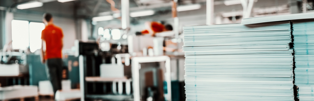 Mehrere Druckbögen aufeinander gestapelt, im Hintergrund Druckereihalle