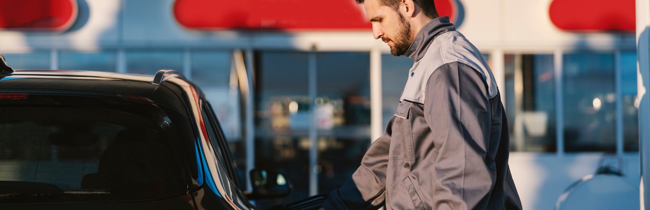 Eine Person in Arbeitskleidung steht an einem Auto auf einer Tankstelle und tankt das Gefährt.