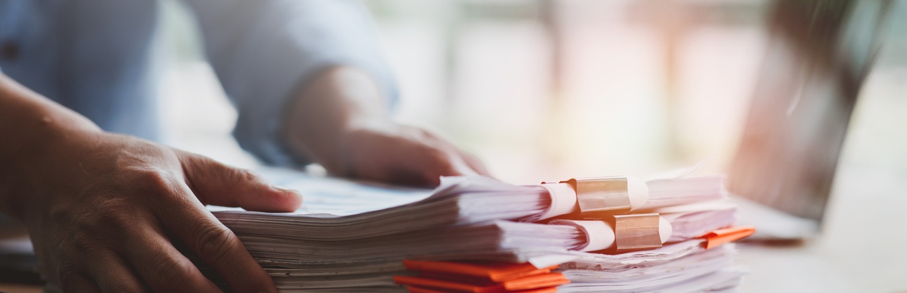Auf einem Tisch liegen mehrere Stapel Papiere. Die Hände einer Person greifen nach dem Stapel