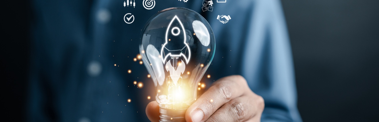 Eine Person hält eine Glühbirne in die Kamera. In der Glühbirne sind die Umrisse einer Rakete. Am unteren Ende ist die Glühbirne beleuchtet. Über der Glühbirne sind grafische Symbole wie eine Zielscheibe, Zahnräder, eine Checkliste oder Puzzleteile