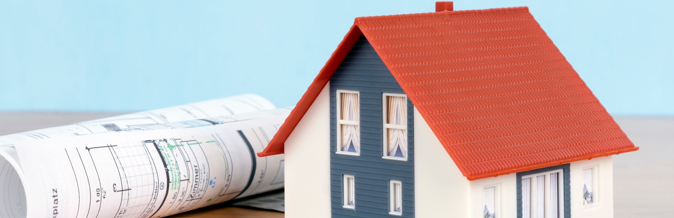 Ein Modellhaus steht auf einer hölzernen Tischplatte. Unter und vor dem Haus liegen verschiedene Eurogeldscheine, auf denen unterschiedliche Zahlen stehen. Neben dem Haus liegt eine Papierrolle, auf der Parkplatz steht. Der Hintergrund ist hellblau.