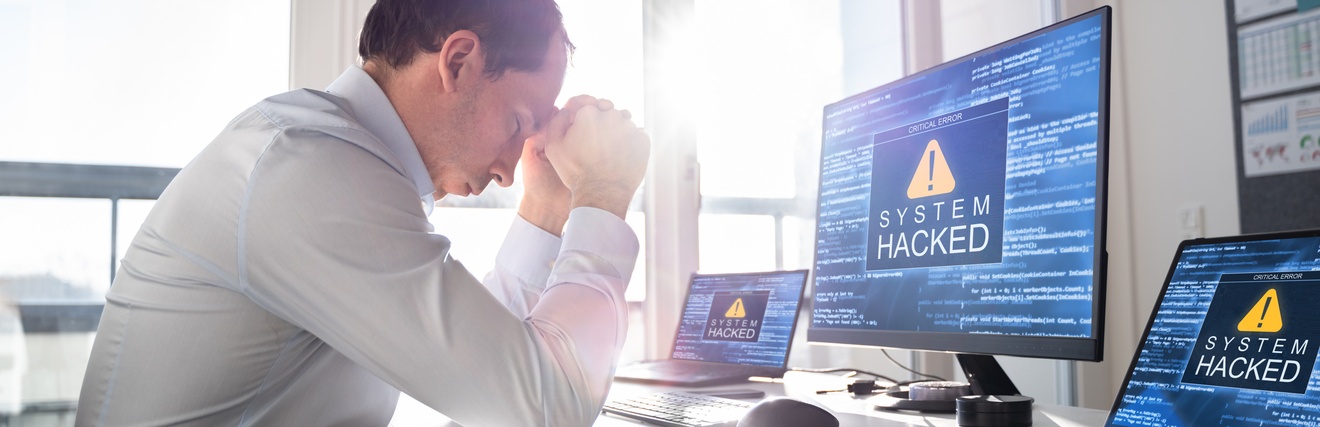 Eine Person sitzt am Arbeitstisch im Büro und hält die Arme auf dem Tisch gestützt vor die Stirn. Auf den Bildschirmen eines Computers und zwei Laptops erscheinen Cyber-Angriff-Symbole mit dem Schriftzug System hacked.