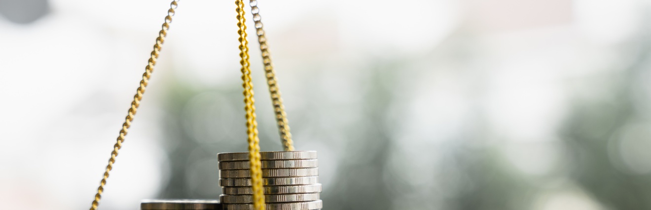 Nahaufnahme einer kleinen, goldenen Schale in der linken Bildhälfte. Am Rand sind drei goldene, kleine Ketten montiert, die oben aufeinander treffen. Auf der Schale liegen drei Münzstapel