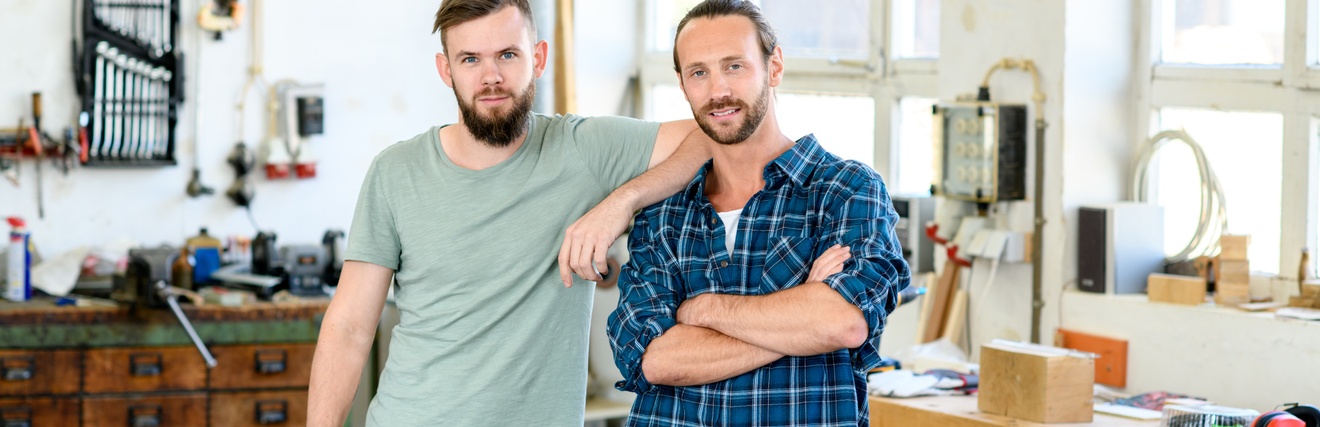Zwei Personen mit Bart stehen zusammen und blicken in die Kamera, im Hintergrund zeigt sich eine Werkstätte