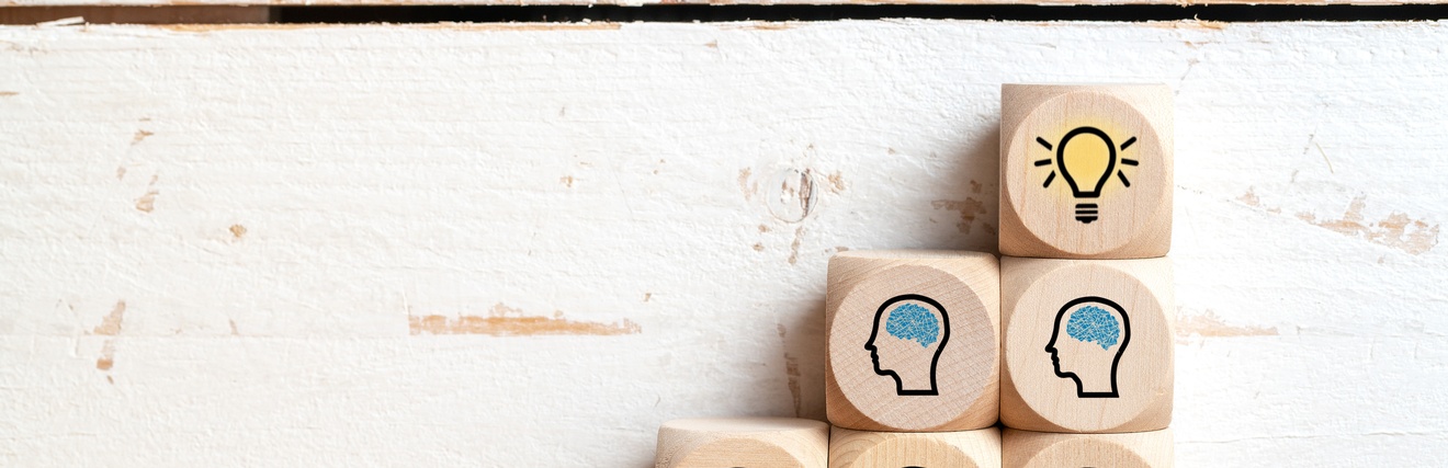 Vor einer hellen Wand stehen fünf Holzwürfel übereinander. Auf fünf Würfeln ist der Umriss eines menschlichen Kopfes, in dem jeweils das Gehirn blau eingezeichnet ist. Auf dem sechsten und obersten Würfel ist eine leuchtende Glühbirne