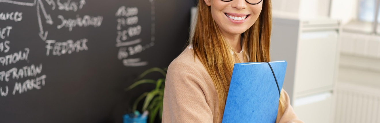 Person mit langen braunen Haaren und Brille hält eine Mappe und steht freudig vor einer Tafel mit Notizen