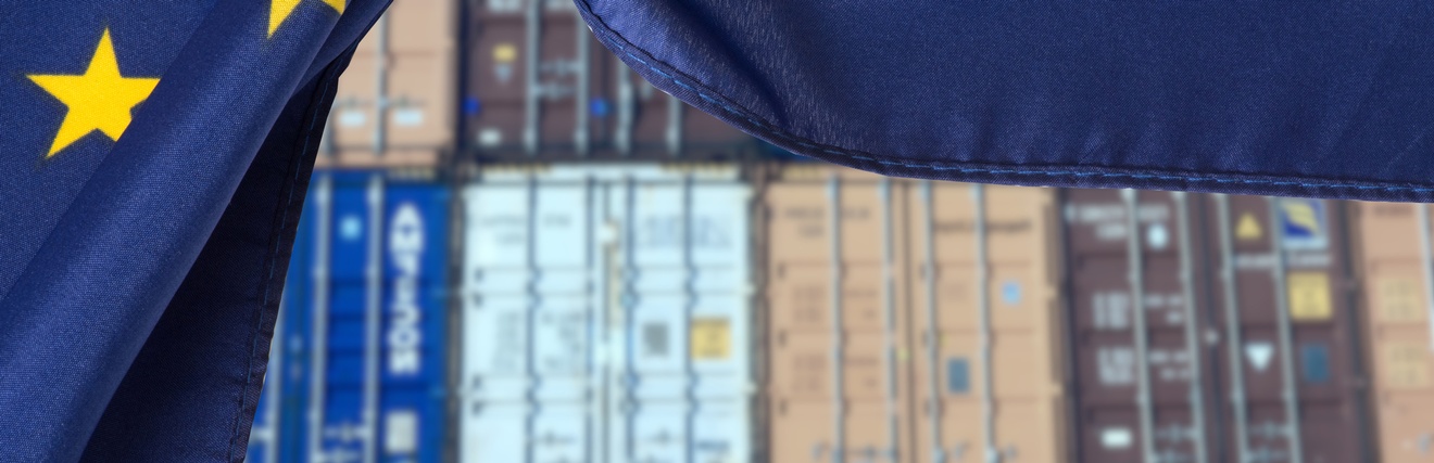 Im Vordergrund ist die Flagge der Europäischen Union, wie ein Vorhand zur Seite geschoben. Dahinter sind gestapelte Container, um Waren zu transportieren