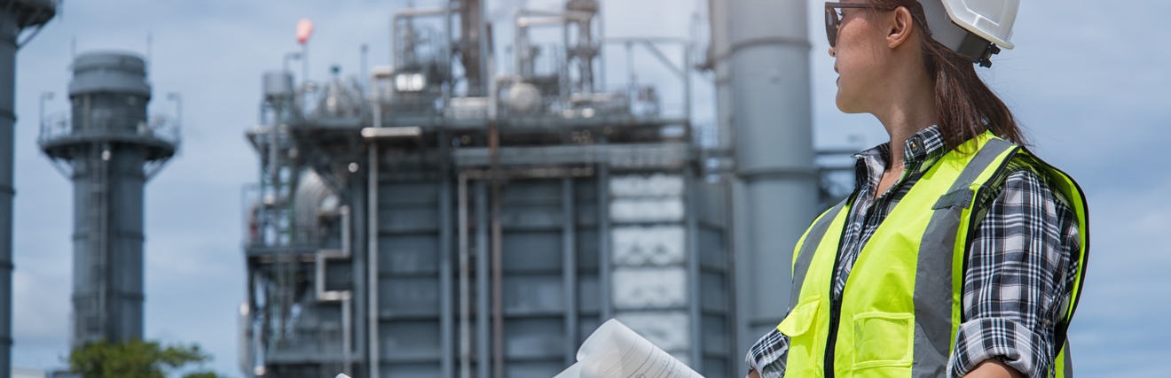 Person mit Warnweste und Schutzhelm hält mit beiden Händen einen Bauplan und blickt zu einem Fabriksgelände