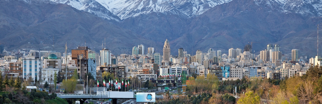 Blick auf die Skyline der Stadt Teheran, gelegen vor dem Elburs-Gebirges mit verschneiten Gipfeln, im Vordergrund sieht man eine befahrene Straße und herbstliche Bäume.