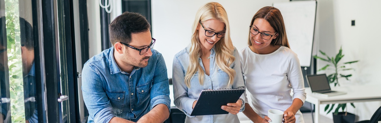 Drei Personen stehen nebeneinander. Die mittlere Person hält einen Notizblock, auf den alle blicken. Im Hintergrund ist ein Whiteboard