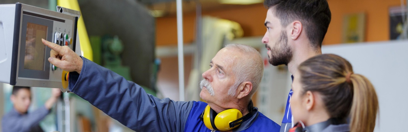 Zwei jüngere Personen in Arbeitskleidung blicken auf Display einer Maschine, auf das ältere Person in Arbeitskleidung mit Hörschutz um den Hals deutet