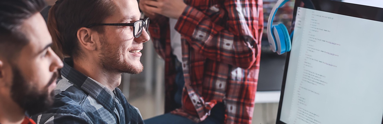 Seitliche Aufnahme zweier neben einem Bildschirm sitzender Personen. Daneben sind zwei weitere stehende Personen. Alle blicken auf den Bildschirm, auf dem mehrere Zeilen sind