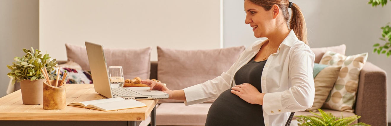 Schwangere Person sitzt freudig bei einem Wohnzimmertisch und arbeitet mit einem Laptop während im Hintergrund eine Couch sowie Pflanzen stehen