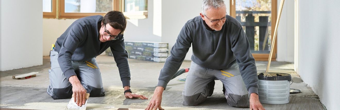 Zwei Personen in grauen Arbeitsmonturen knien am Boden und verlegen Holzbodenbretter, im Hintergrund Fenster, Balkontüre und Fernseher
