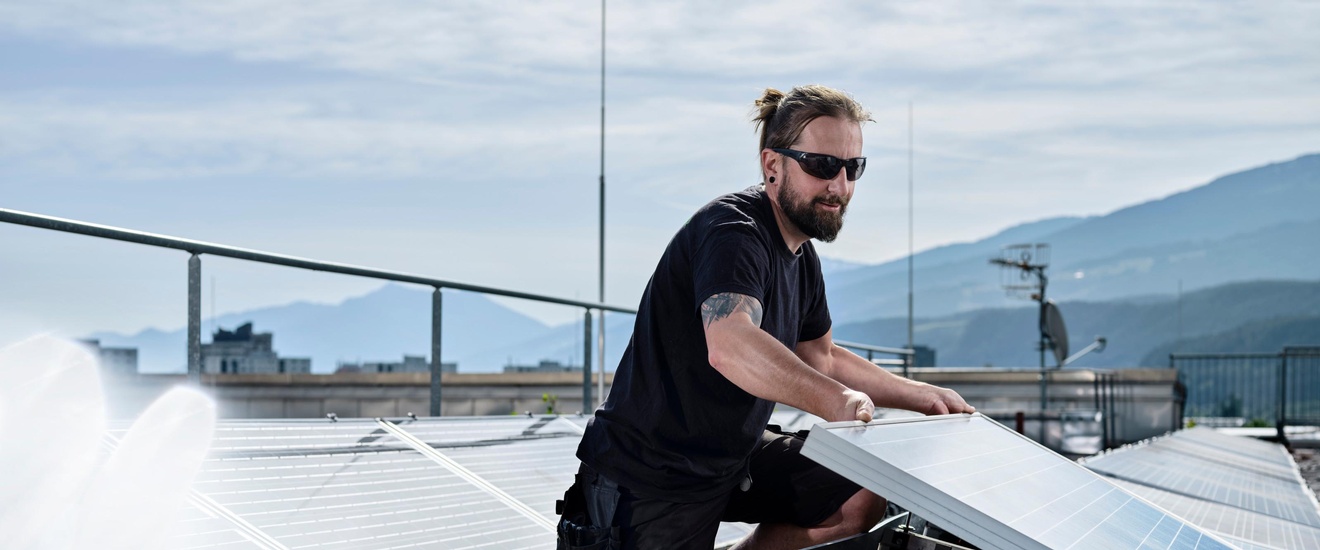 Ein Mann mit Sonnenbrille installiert Photovoltaikanlagen auf einem Flachdach. Im Hintergrund bergige Landschaften.