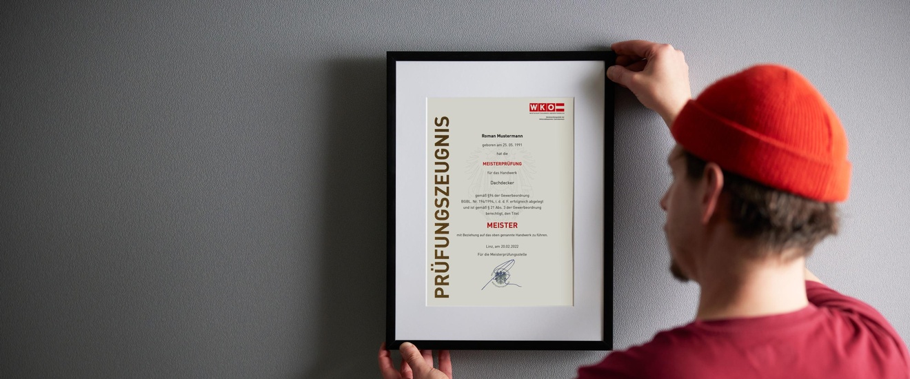 Person mit kurzen dunklen Haaren, Bart, roter Mütze und rotem Shirt steht vor einer grauen Wand und hält einen Bilderrahmen mit Passepartout und Prüfungszeugnis Meister in Händen, daneben steht eine Pflanze