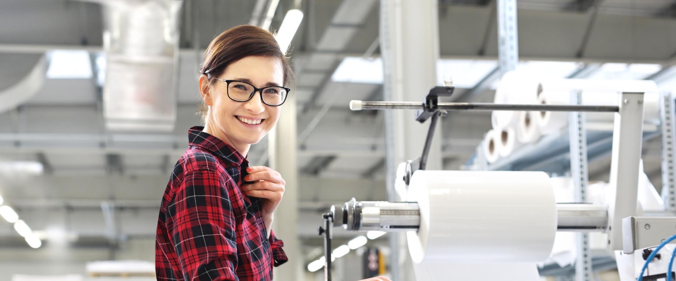 Eine Person mit Brille steht seitlich vor einer großen Maschine. Auf einer Fläche der Maschine liegt ein hoher, weißer Papierstapel. Schräg über dem Stapel ist eine Metallrolle, auf der eine dicke, weiße Rolle ist. Die Person blickt direkt in die Kamera.