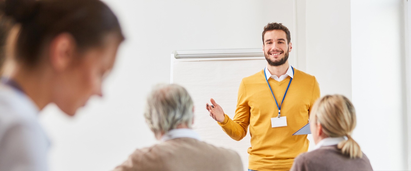Rückenansicht von fünf Personen, die in einem Raum an Tisch sitzen. Zwei Personen haben vor sich einen aufgeklappten Laptop. Ihnen gegenüber steht eine Person, die einen Ausweis um den Hals baumeln hat und ein Klemmbrett hält.