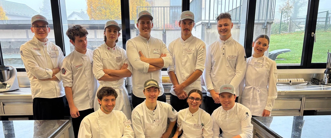 Porträt von insgesamt elf jungen Personen, die in zwei Reihen in einer Gastronomieküche in Kochkleidung stehen und lächeln