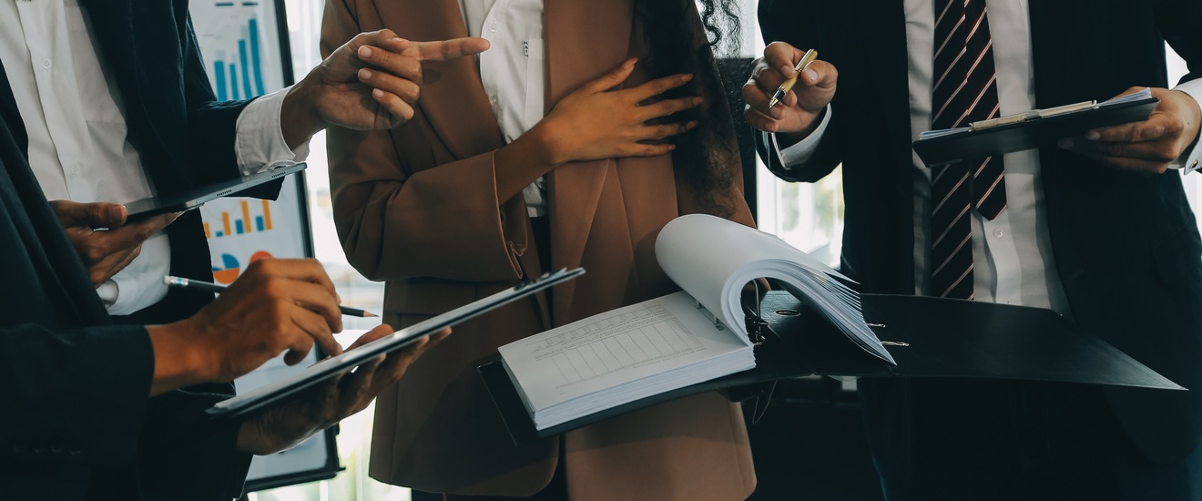 Aufnahme der Oberkörper von vier Personen in Bürokleidung. Zwei Personen halten ein Klemmbrett, eine Person ein Tablet, eine Person einen aufgeklappten Ordner mit vielen Zetteln. Im Hintergrund ist eine Tafel mit Statistiken