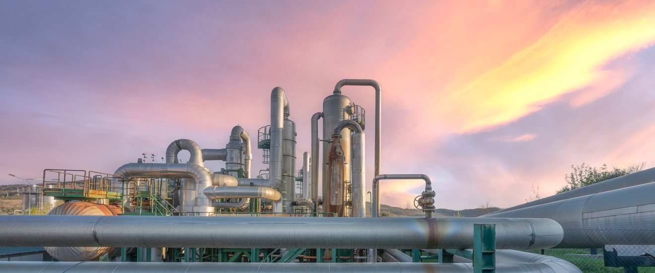 Vor rötlichem Himmel verlaufen im Vordergrund Pipelines. Dahinter sind weitere Pipelines sowie Silos und Metalltreppen. Rechts ist eine grüne Wiese