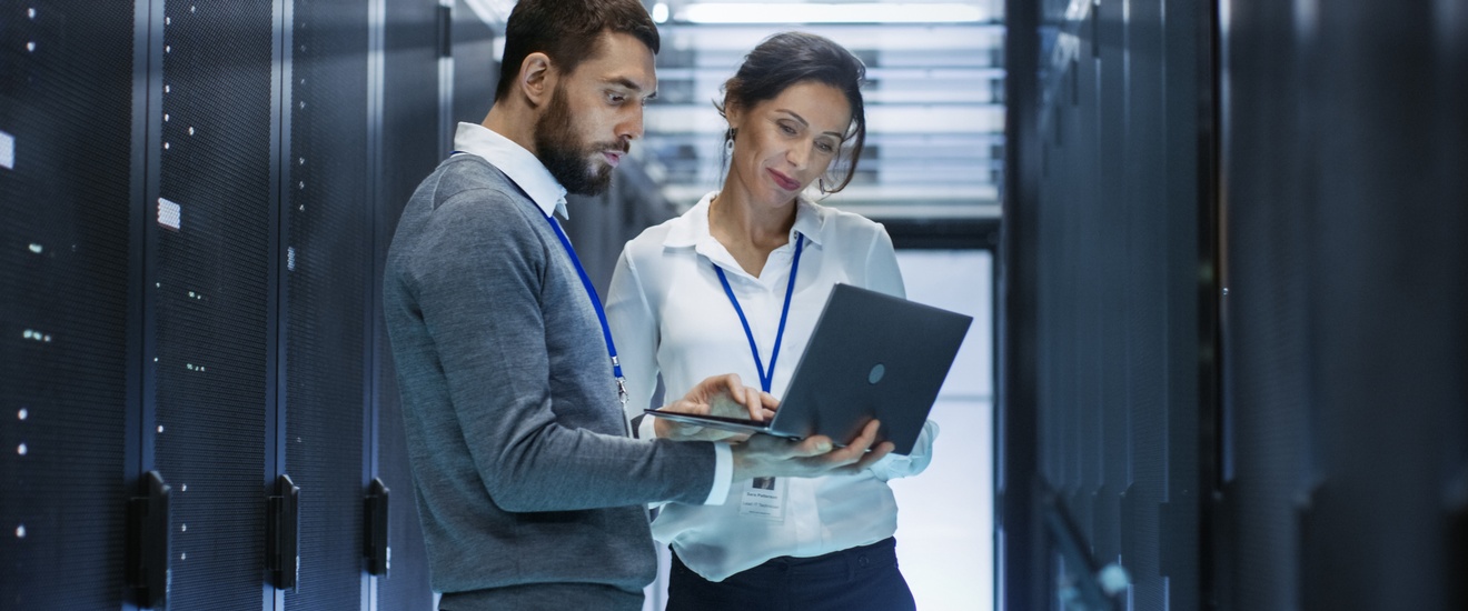 Zwei Personen stehen vor Servern und blicken auf aufgeklapptes Notebook, das eine Person in Händen hält